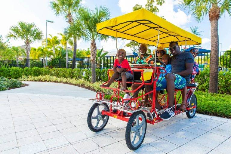 A multigenerational family of grandparents, parents, and children ride on a Surrey bike around the Encore Resort at Reunion.