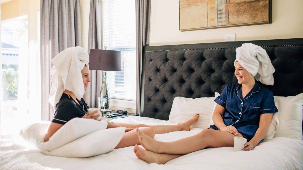 अपने बालों पर नहाने के तौलिये के साथ दो महिलाएँ एक शाही बिस्तर पर बैठी और बातें कर रही हैं Encore Resort लड़कियों के पलायन के दौरान निवास।