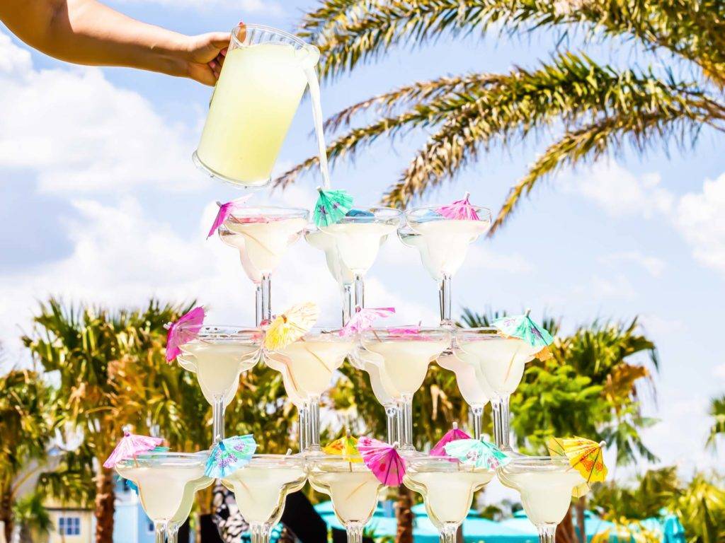 Margaritas being poured from a single pitcher into a stacked tower of glasses.