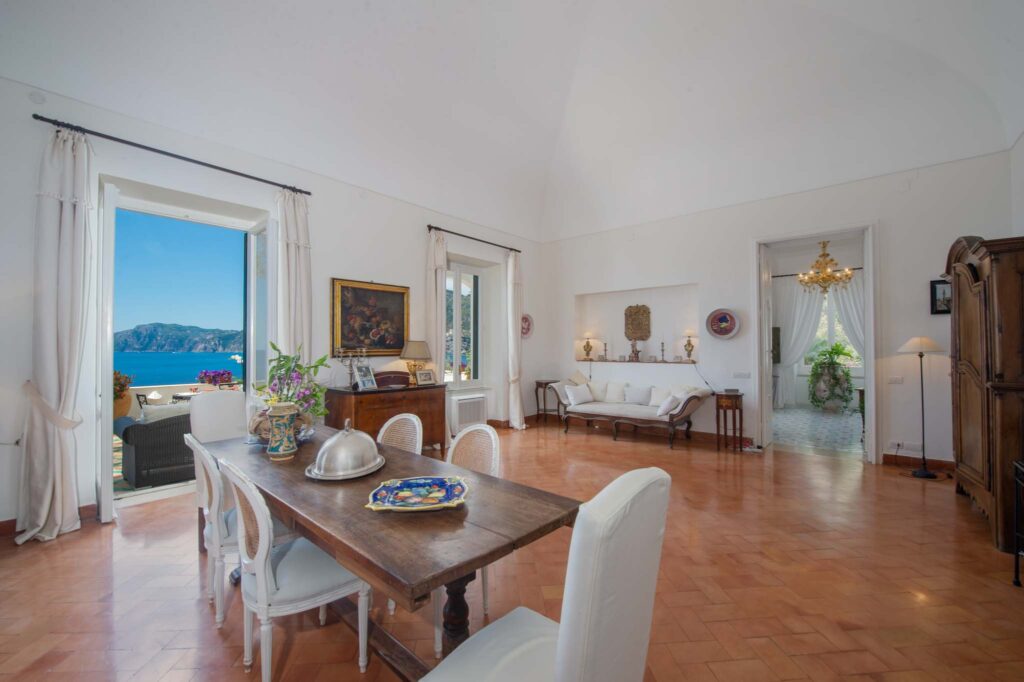 Juego de mesa de comedor para 6 personas en la espaciosa sala de estar de Villa Lilly con una gran ventana que ofrece una vista impresionante del mar Tirreno y la costa de Amalfi.