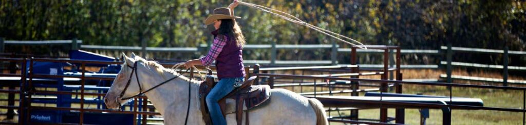 Woman riding horseback swinging a rope lasso at The Ranches at Belt Creek rodeo grounds.