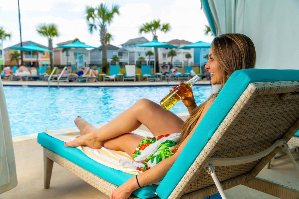 امرأة مبتسمة تجلس تحت كابانا خاصة مع زجاجة من بيرة لاند شارك بجوار مسبح Margaritaville Resort Orlando.