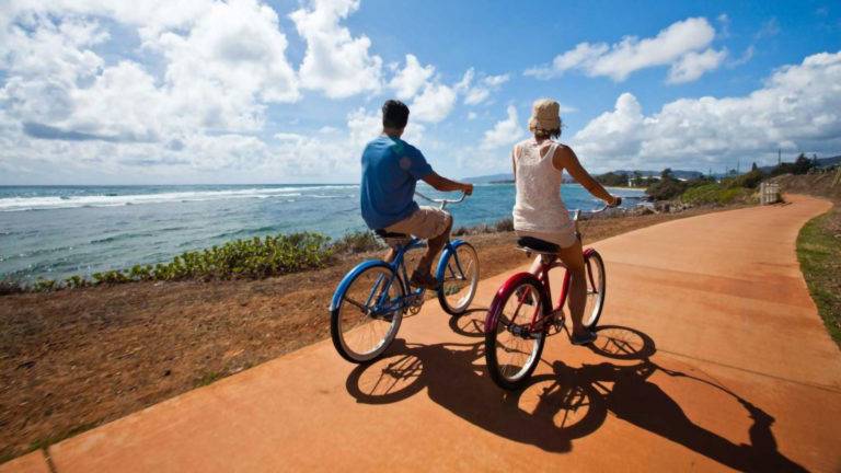 Two people riding bikes on a bicycle trail by Pono Kai Resort next to the ocean.