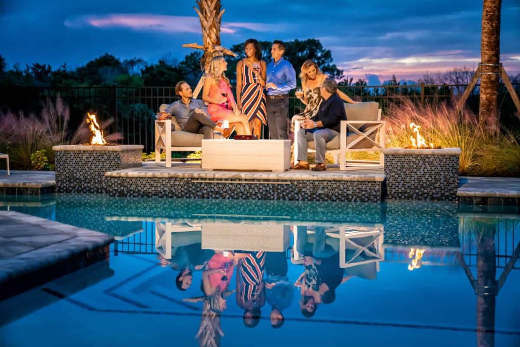 Grupo de parejas sentadas y conversando junto a un par de fogatas junto a la piscina en Spectrum Resort Orlando al atardecer.