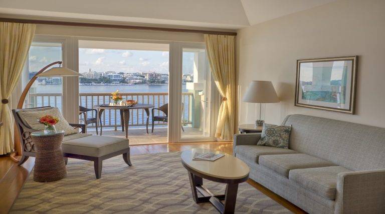 جناح غرفة المعيشة مع أريكة وكرسي قراءة وشرفة خاصة تطل على ميناء هاميلتون في برمودا Rentyl Resorts.