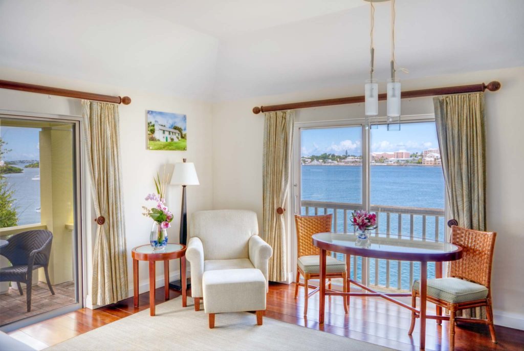 Habitación tipo estudio de lujo con silla de lectura, mesa de comedor pequeña y balcón privado con vista al puerto de Hamilton en las Bermudas Rentyl Resorts orlando