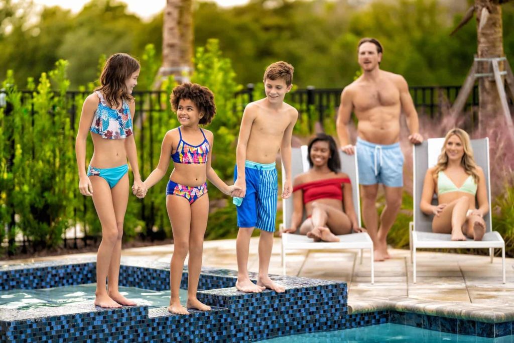 Niños tomados de la mano en el borde de una piscina privada mientras los padres los ven jugar felices.