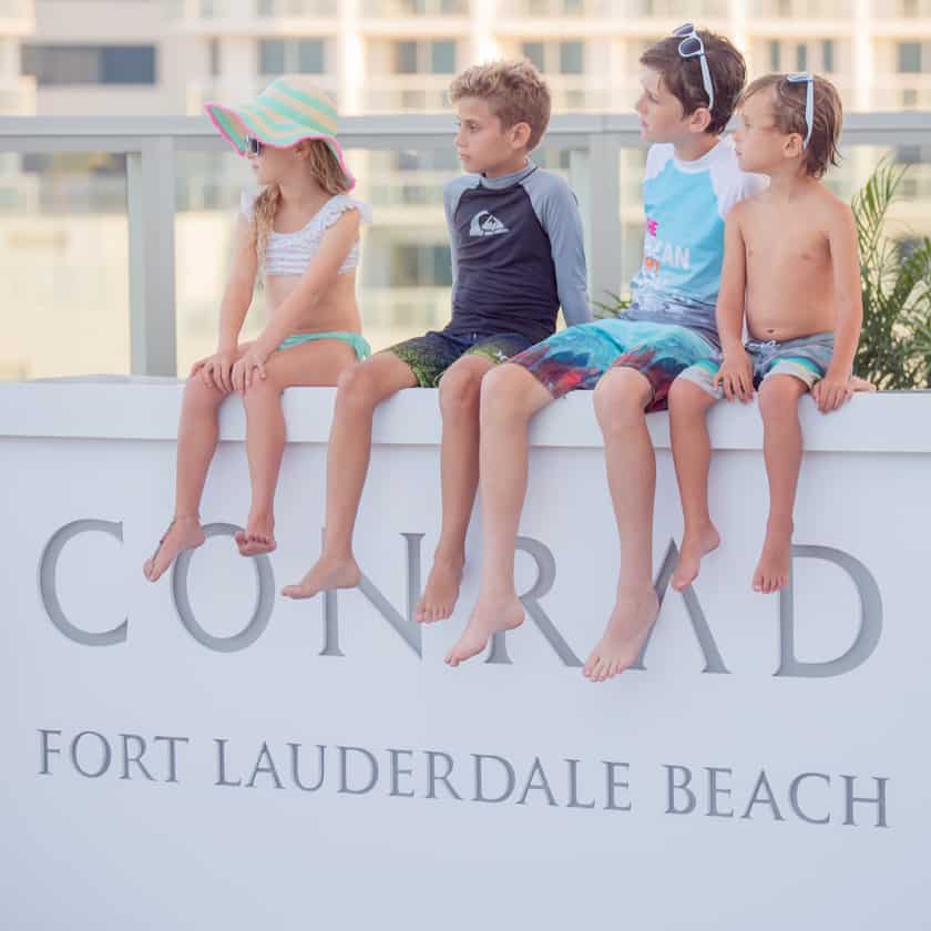 Kids in swim suits sitting on the Conrad Fort Lauderdale Beach sign