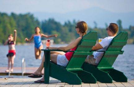 एडिरोंडैक कुर्सियों पर बैठे माता-पिता बच्चों को झील में कूदते देख रहे हैं
