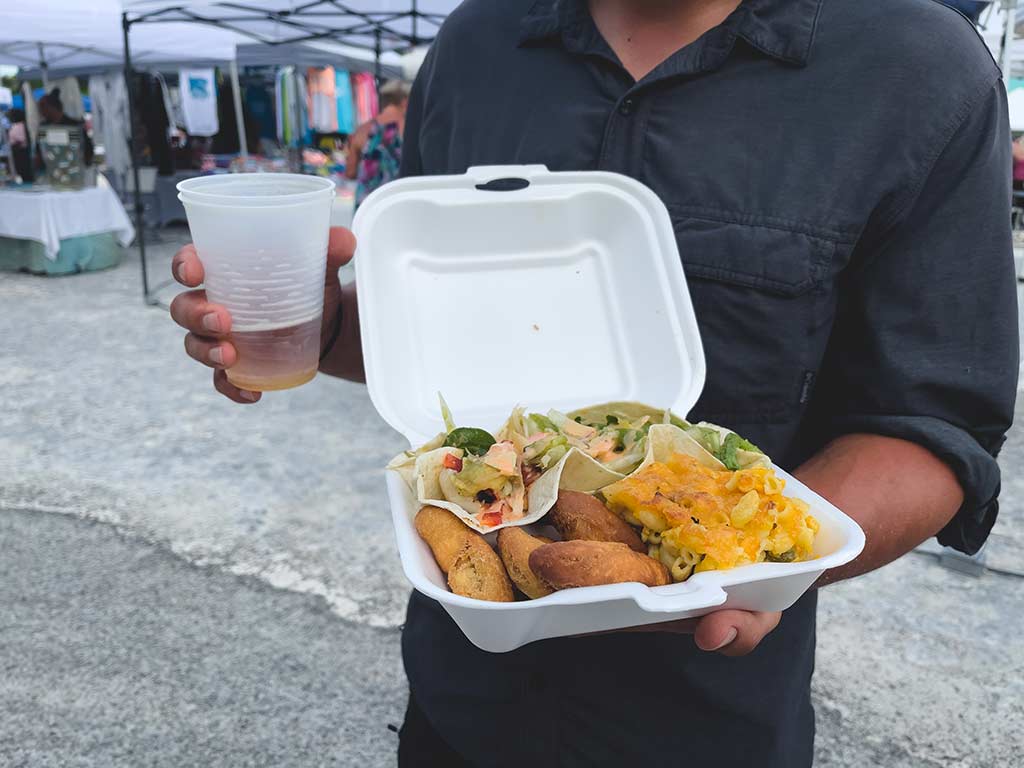 Man holding a take out container with a variety of food and a beer.