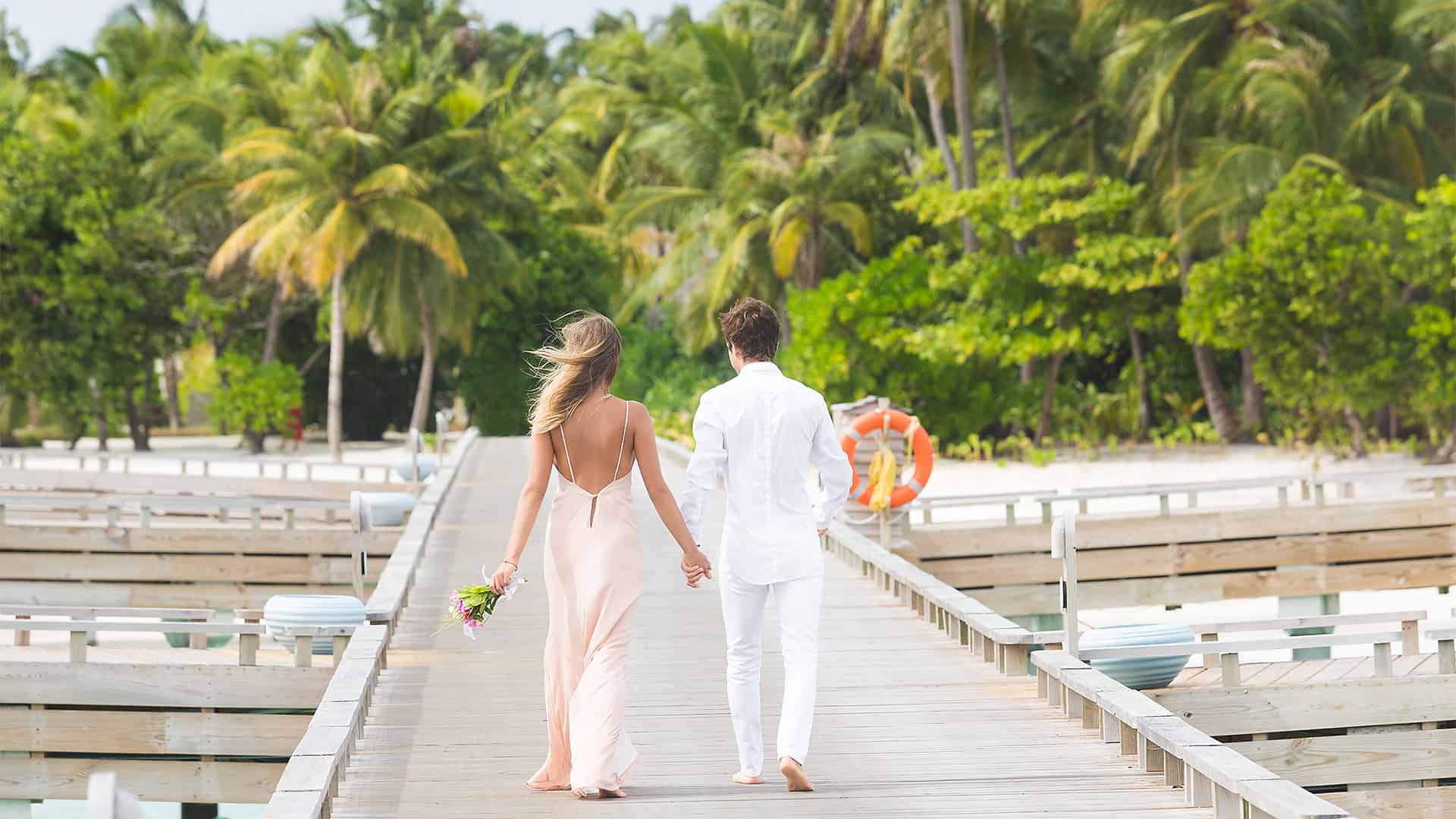 La novia y el novio caminan de la mano por un pasillo de bodas en la playa con palmeras.