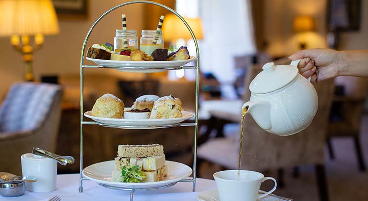 Person pouring a cup of tea next to a tiered serving tray with sandwiches, biscuits, and desserts for Afternoon Tea at the Greenway Hotel