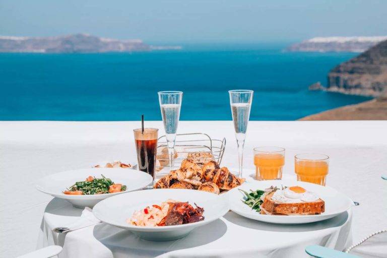 Platos, pasteles, jugos y champaña en una mesa de comedor al aire libre con vista al mar para las cenas en la habitación de Athina Luxury Suites