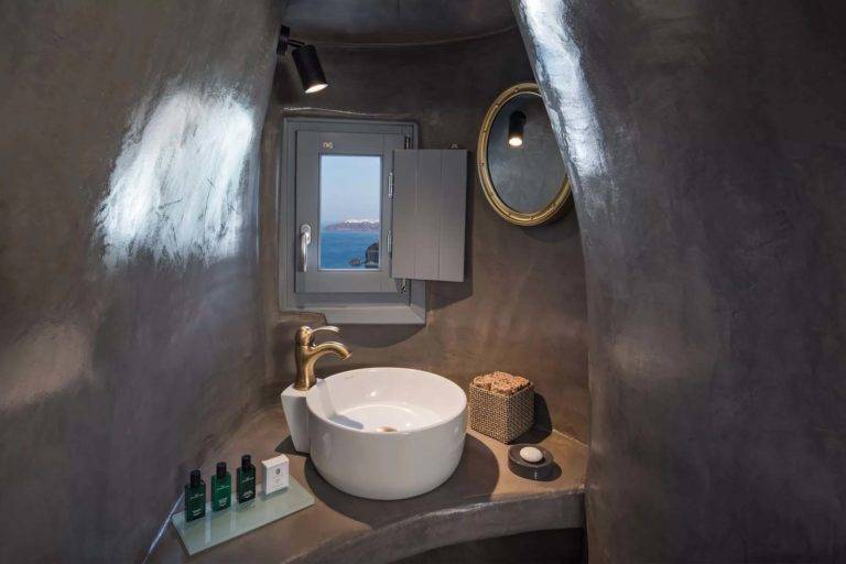 Suite Petit Cave - Baño con tocador, espejo, artículos de tocador y ventana pequeña con vista al mar | Suites de lujo Athina