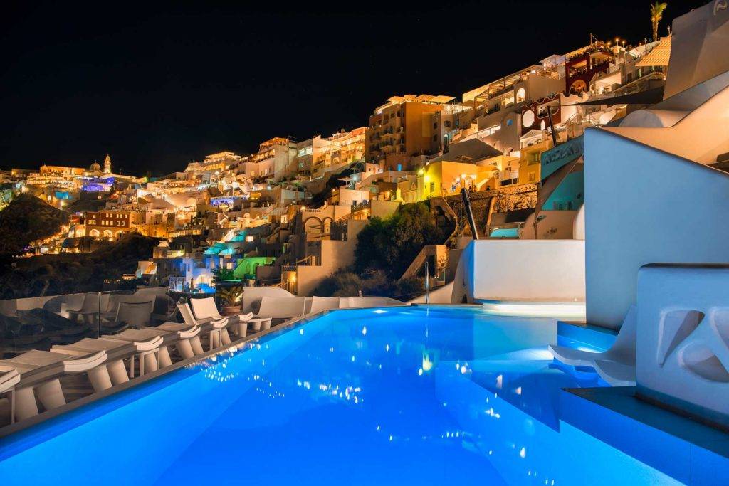 Piscina infinita de Athina Luxury Suites iluminada por la noche con vistas a la ciudad de Fira