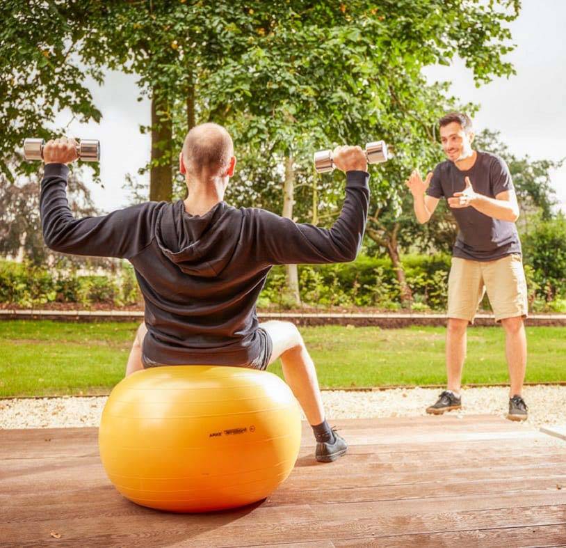 Hombre levantando pesas sentado en un balón medicinal y trabajando con un entrenador personal en Mallory Court