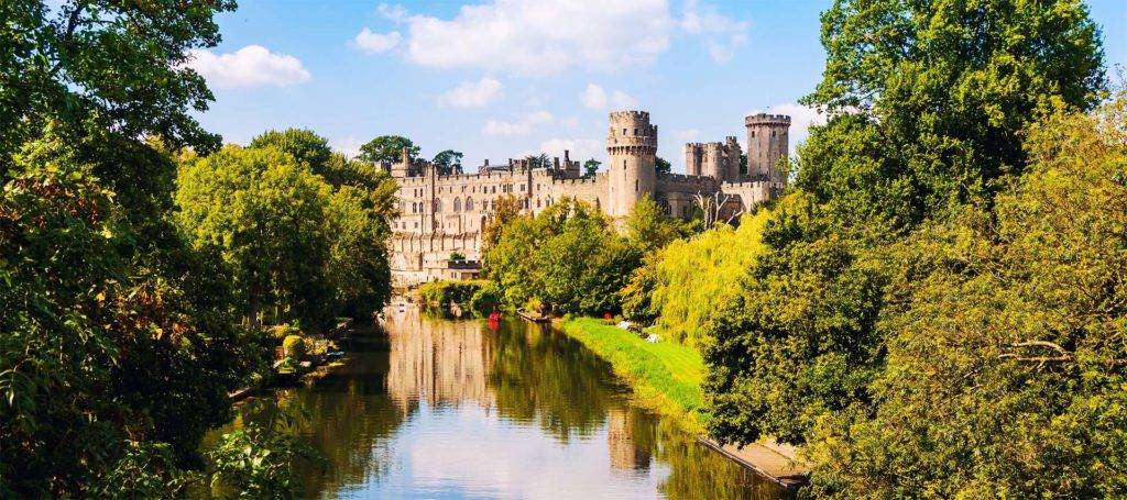 Exterior view of Warwick Castle surrounded by greenery and a river in Warwickshire, United Kingdom