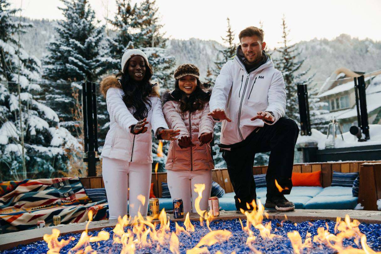 Dreiergruppe in Winterkleidung, die sich die Hände auf der Feuerstelle auf der Dachterrasse des W Aspen in Colorado wärmt
