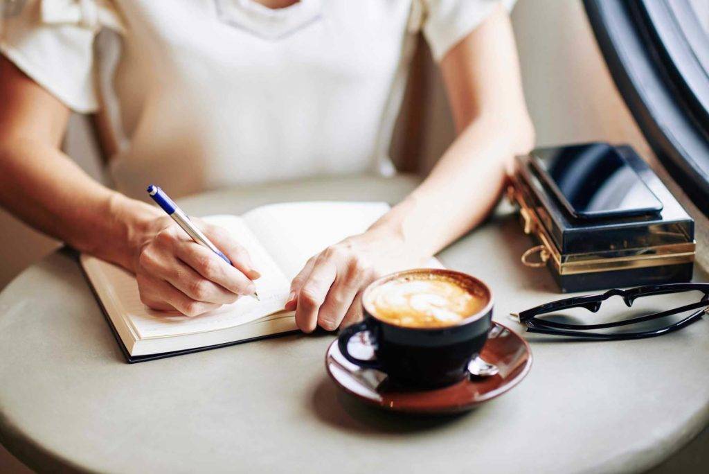 Mujer escribiendo en un diario en una cafetería mientras bebe un café con leche