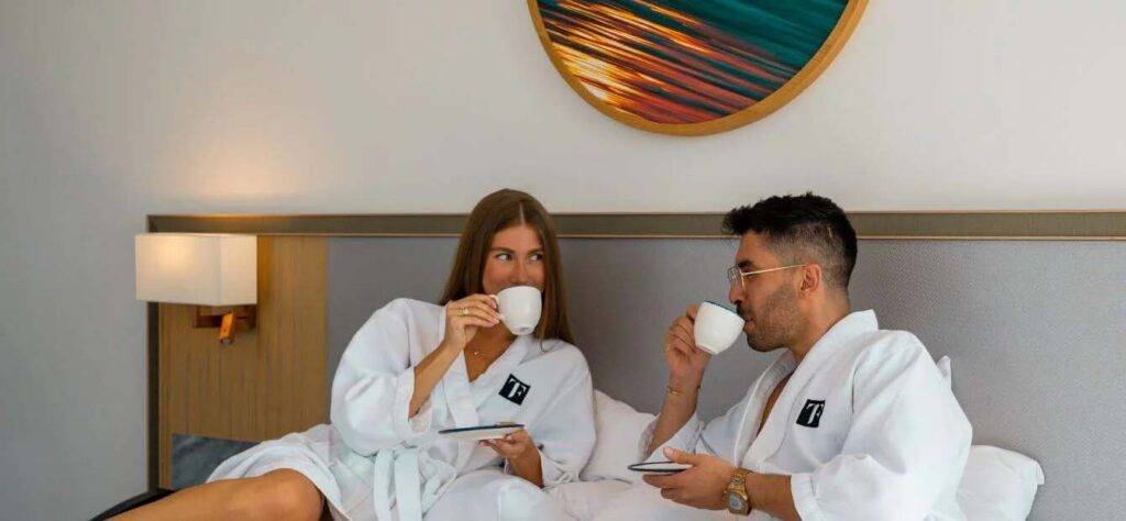 Ein Paar entspannt sich in Bademänteln und trinkt Kaffee im Bett seines First Collection JVC Hotelzimmers