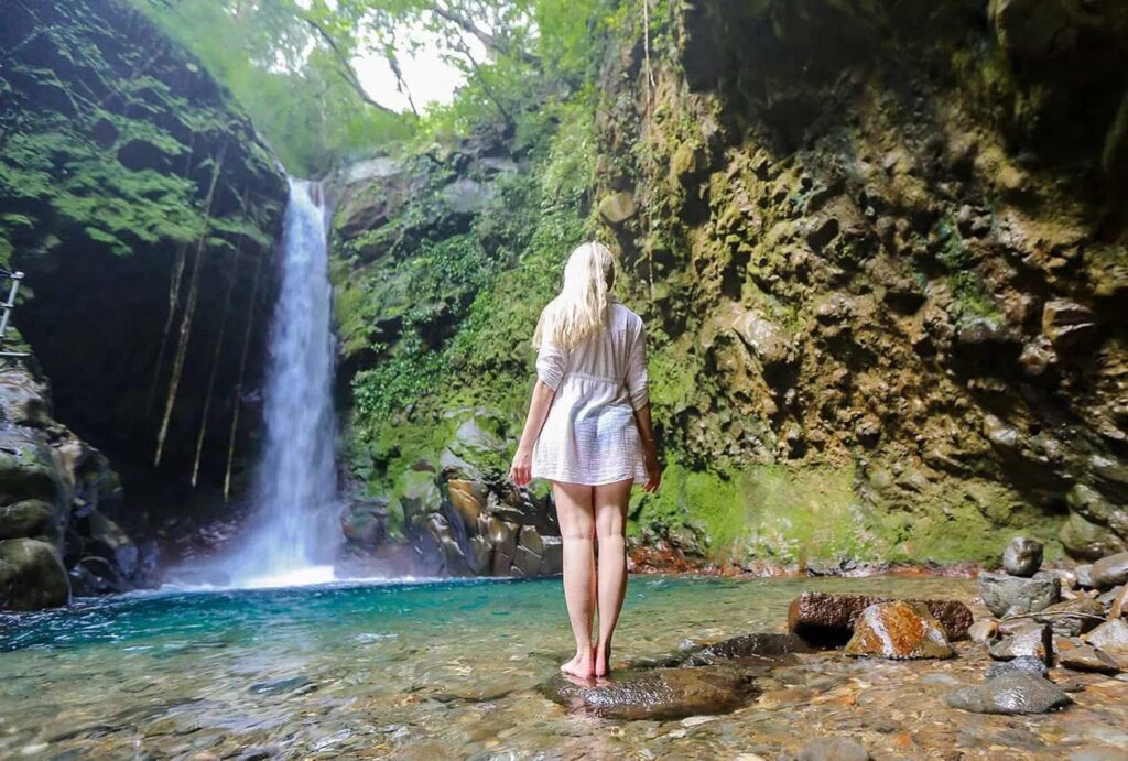 Mujer sumergiendo sus pies en un río mientras admira una cascada en la selva tropical de Costa Rica