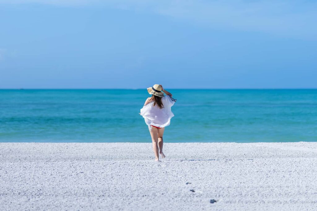 सन हैट और स्विमसूट पहने महिला द रिज़ॉर्ट एट लॉन्गबोट की क्लब में मेक्सिको की खाड़ी की ओर साफ़ रेतीले समुद्र तट पर टहलती हुई